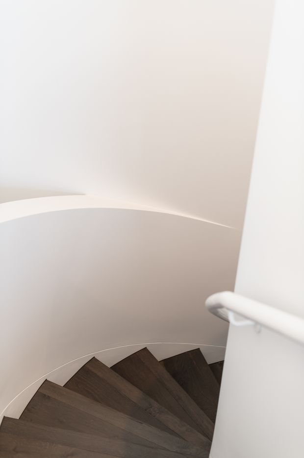 A minimalist curved stairway viewed from above, featuring dark timber stair treads winding around a central curved wall. The surrounding walls and ceiling are smooth white plaster, with a simple white handrail following the stair curve.