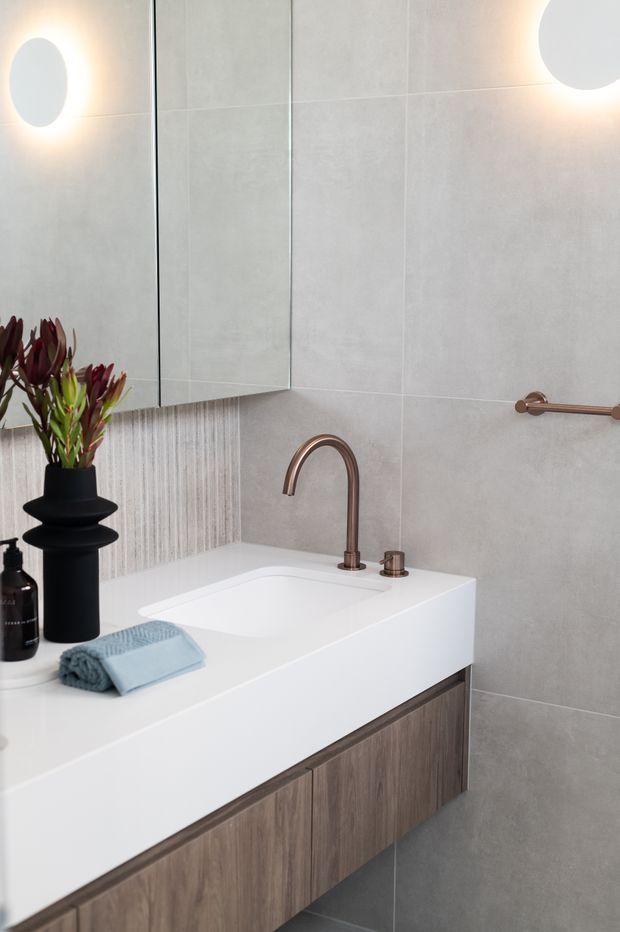 Contemporary bathroom vanity with a white integrated basin, warm metallic curved tapware, wood-grain floating cabinetry, large pale grey wall tiles, and a mirrored cabinet. Soft wall lighting, a folded blue towel, and a sculptural dark vase create a refined, calm fit-out.