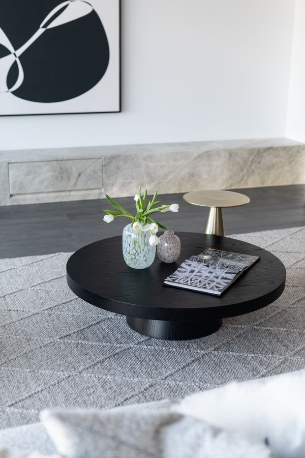A refined contemporary living room vignette featuring a large round black coffee table with a pedestal base, a brass-toned side table, textured grey rug, pale stone or marble fireplace surround, and minimalist black-and-white artwork. Decorative vases and a magazine sit on the coffee table, with a soft neutral upholstered sofa partially visible in the foreground.