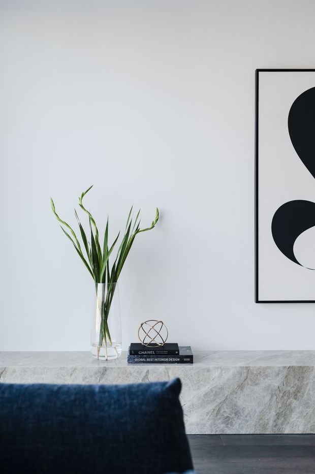 A refined interior vignette featuring a pale grey wall, a marble-clad console or low built-in surface, a clear glass vase with tall green foliage, stacked design books, and a small geometric sculpture. A large black-and-white abstract artwork hangs above, while the foreground includes part of a deep navy upholstered chair or sofa. Dark timber flooring and restrained styling create a calm, contemporary atmosphere.