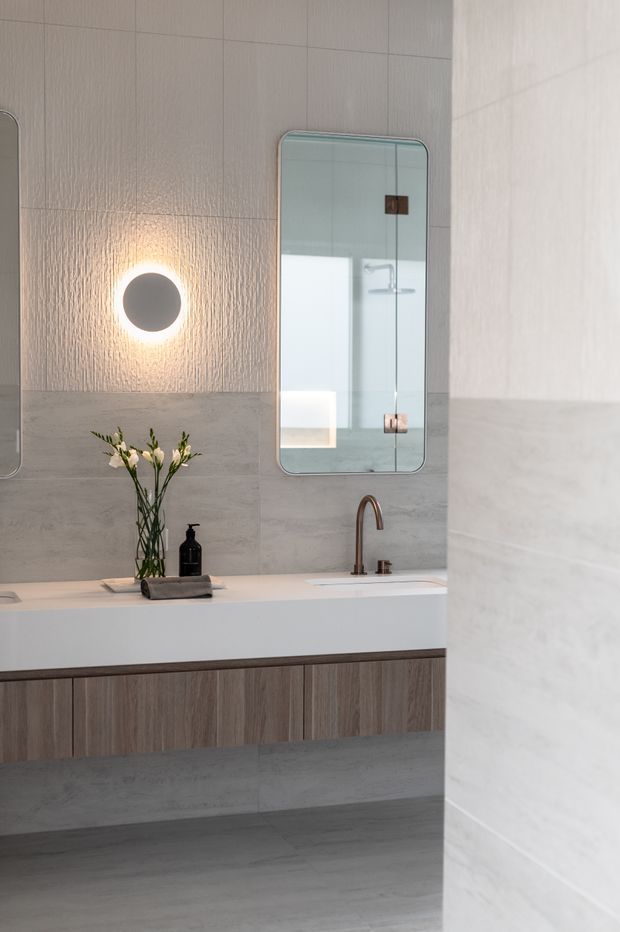 A refined contemporary bathroom featuring a floating double vanity with warm wood-grain cabinetry, a pale stone or porcelain benchtop, integrated basin, and brushed bronze tapware. Large grey-beige stone-look wall and floor tiles create a calm, tonal backdrop, complemented by rounded-rectangle mirrors and a glowing circular wall light. Minimal styling includes a vase of flowers and a dark soap dispenser.