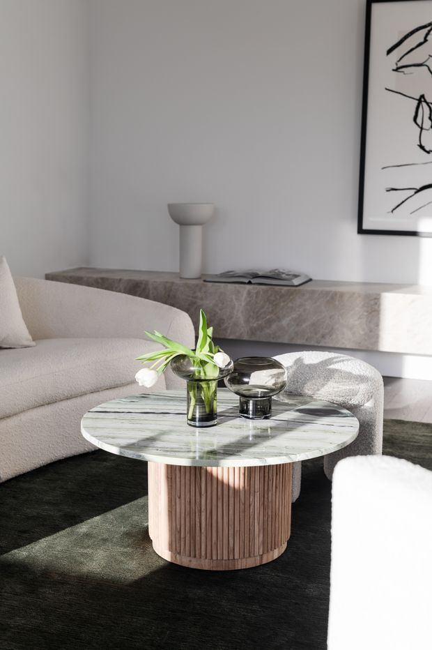A bright, refined contemporary living room featuring a curved cream boucle sofa, a round coffee table with a pale veined stone top and vertically fluted timber base, dark textured flooring, and sculptural glass vessels with greenery. A long stone-clad console runs along the rear wall beneath minimalist black-and-white artwork, with sunlight creating strong highlights and shadows.