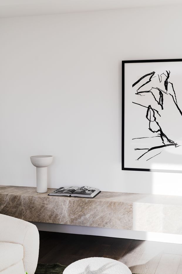 A minimalist interior vignette featuring a long floating stone console with a veined beige-grey finish, a white sculptural vase, and an open book. A black-framed abstract line artwork hangs above against a white wall, with partial white upholstered seating and dark timber flooring visible in the foreground. Strong natural light creates a calm, refined atmosphere.