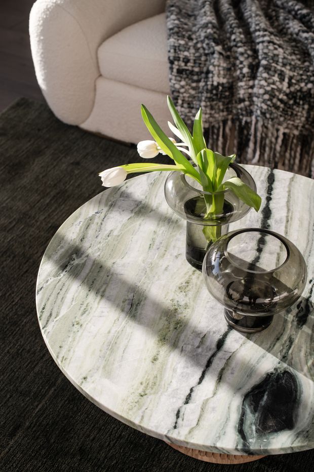 Close-up interior vignette showing a round polished natural-stone coffee table with dramatic white, grey, and green veining, set on a dark textured rug. Two smoky glass vessels and a clear glass vase with white tulips sit on the table, while a cream boucle armchair and chunky black-and-white knitted throw are visible behind it.