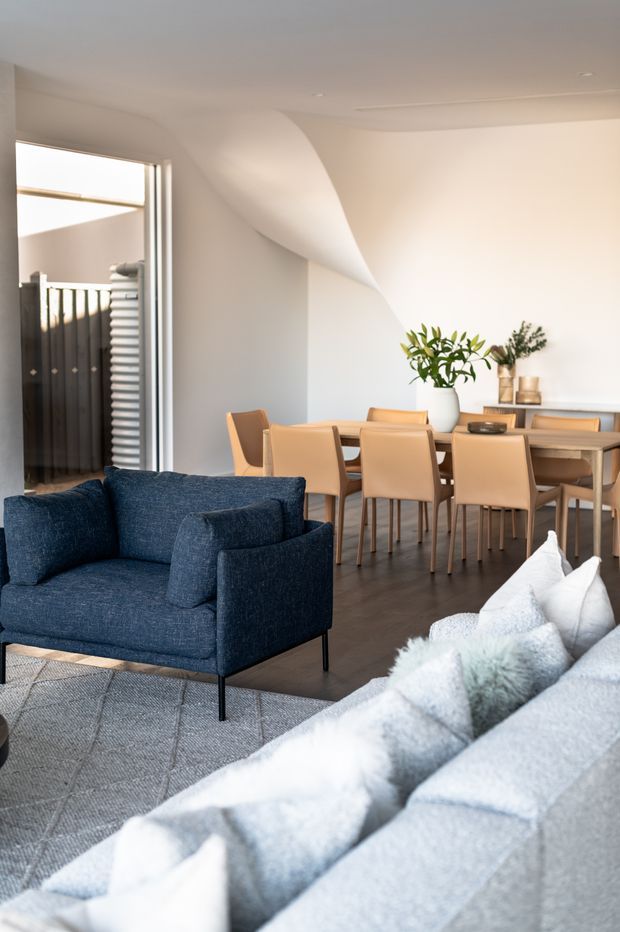 A bright, refined open-plan living and dining area. A deep navy-blue sofa and pale grey sectional frame a timber dining table with tan upholstered chairs. The room has warm light walls, dark timber flooring, recessed ceiling lighting, large glazed opening, and a sculptural curved ceiling-wall detail. Minimal decor includes a white vase with greenery and a low sideboard.