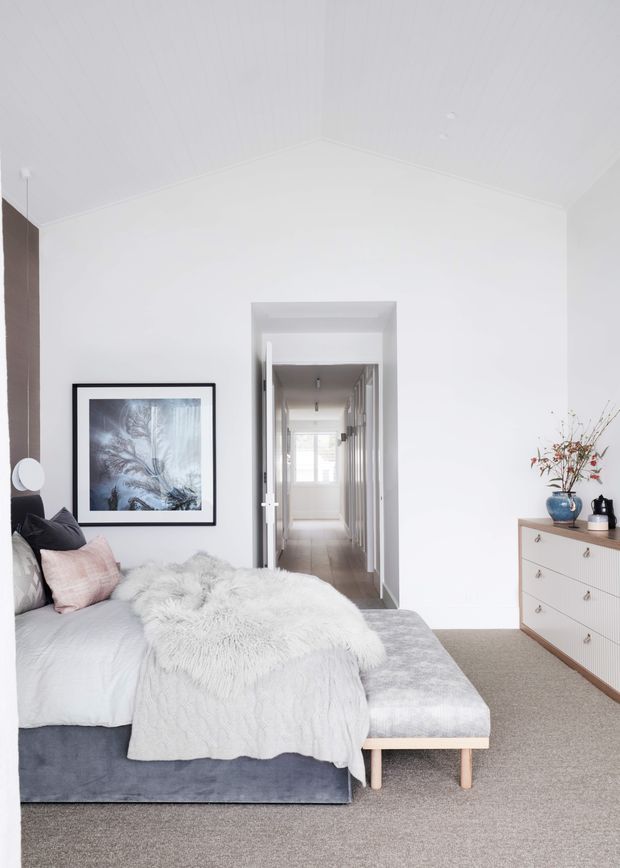 Bright, serene bedroom with a low upholstered bed dressed in pale grey and white bedding, layered fluffy throws and cushions, a light upholstered bench at the foot, and a simple black-framed artwork. White walls and a vaulted white ceiling create an airy backdrop, while a pale wood dresser with a ribbed front and delicate hardware sits beside a blue ceramic vase and dried branches. An open doorway leads to a bright hallway.