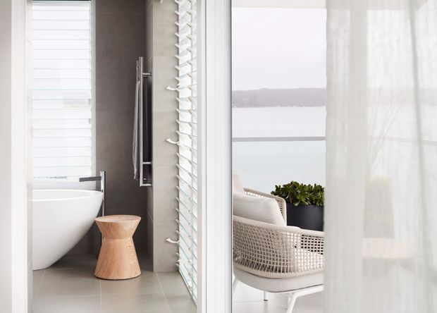 Bright contemporary bathroom with a freestanding white bathtub, wall-mounted fixtures, grey tiled surfaces, and a sculptural timber stool. Through the adjacent glazing is a covered outdoor seating area overlooking calm water, with woven furniture and a planter.