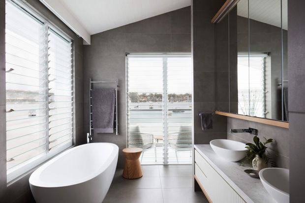 Contemporary bathroom with a freestanding white oval bathtub, double vanity with vessel basins, dark grey wall tiles, pale floor tiles, large mirrored joinery, heated towel rail, and plantation-shuttered windows. Water is visible beyond the glazing, providing a bright waterfront outlook.