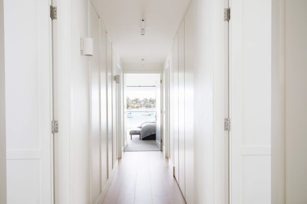 Bright, minimalist white hallway lined with full-height built-in cabinetry and multiple door openings, leading toward a bedroom and a large window with a glimpse of water and buildings beyond. The corridor has pale timber flooring, white walls and doors, simple cylindrical wall lights, and recessed ceiling lighting.