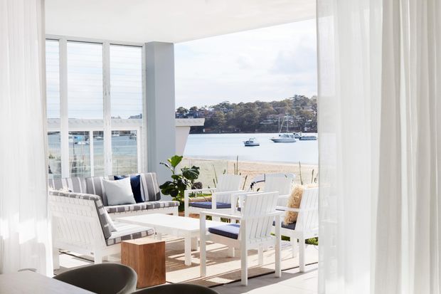 Bright coastal living area opening onto a waterfront terrace, furnished with white outdoor-style lounge seating, a coffee table, and dining chairs. Floor-to-ceiling glazing and sheer white curtains frame views across the water, creating an airy indoor-outdoor setting.