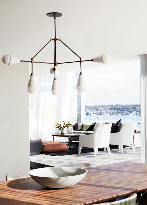 Bright, refined open-plan living and dining area with expansive waterfront glazing. The room features a large sculptural multi-arm pendant light, white lounge chairs, a dark sofa, a low round coffee table, timber dining table, and neutral contemporary furnishings. A marina and residential shoreline are visible through the windows.