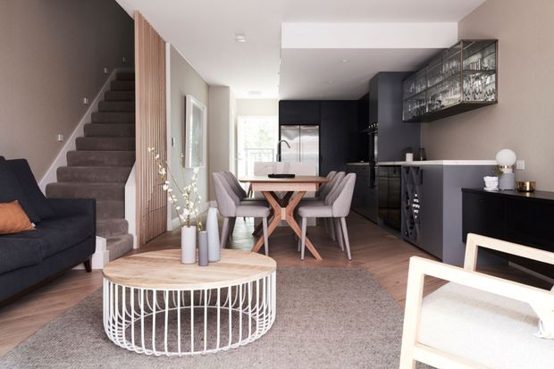 Contemporary open-plan living and dining area with a dark kitchen at the rear. The room features warm timber flooring, a carpeted staircase, vertical timber screening, a timber dining table with upholstered chairs, a dark sofa, a round white wire-frame coffee table, and a wall-mounted glass-front display cabinet. The space is bright with a neutral palette and strong charcoal accents.