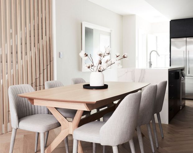 Bright contemporary open-plan dining area with a large light timber dining table and upholstered dining chairs. A vertical timber batten screen forms a feature wall on the left, while a white marble-look kitchen island, stainless-steel appliances, and pale neutral finishes are visible beyond.