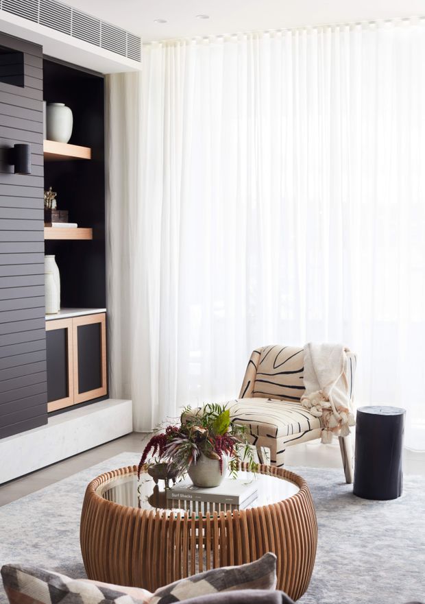 Bright contemporary living room with sheer floor-to-ceiling curtains, a dark built-in shelving and media wall with timber shelves and lower cabinetry, a sculptural upholstered armchair, round slatted timber coffee table, and neutral soft furnishings. The room has abundant natural light and a refined, layered neutral palette.