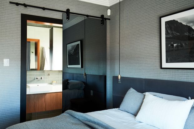 A contemporary bedroom with a low upholstered dark headboard, white bedding, and a large black-framed monochrome artwork. The room features textured grey wallcovering, black-framed sliding barn-style doors, and warm timber bathroom cabinetry visible through the opening. A compact bathroom with a white basin and tiled surfaces is clearly connected directly to the bedroom.