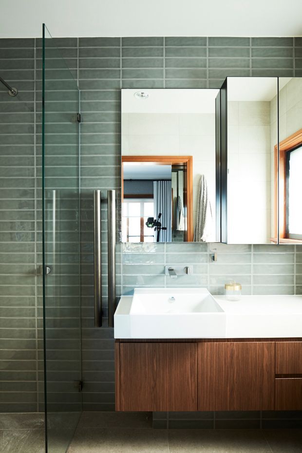 Contemporary bathroom featuring a walk-in glass shower, grey-green rectangular wall tiles, a white vessel-style basin, mirrored wall cabinets, and a floating dark timber vanity with a white benchtop. Warm timber-framed window detailing and brushed metal fixtures add contrast.