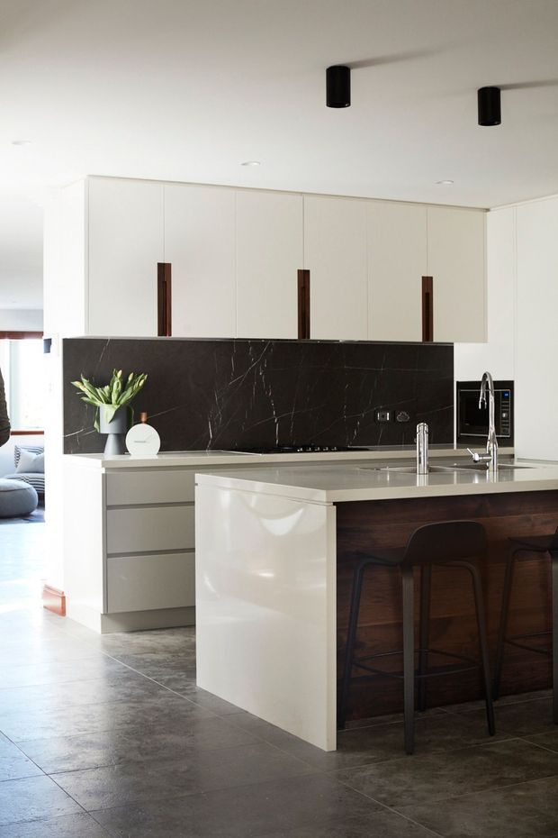 Bright contemporary kitchen with white full-height cabinetry, dark marble-look splashback, pale stone island benchtop, integrated appliances, and a dark timber island face with black bar stools. Large-format dark grey floor tiles and minimalist black cylindrical ceiling lights complete the clean, refined space.