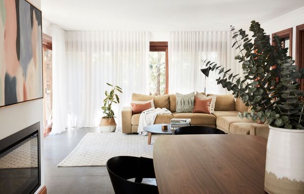 Bright, contemporary living room with a large tan sectional sofa, round black coffee table, curved wood dining table in the foreground, sheer floor-to-ceiling curtains, a modern fireplace with abstract artwork, and large indoor plants. The room has a warm neutral palette and abundant natural light.