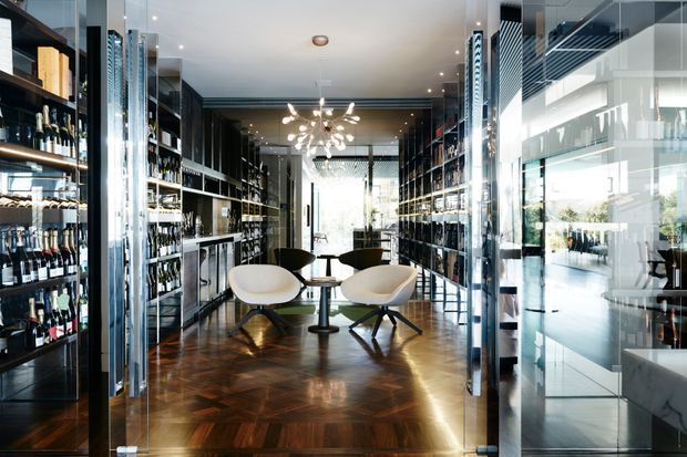 A sophisticated wine bar or retail showroom with floor-to-ceiling glass partitions, extensive dark shelving displaying wine bottles, a central seating area with sculptural lounge chairs and a small round table, and a polished parquet timber floor. Reflective metal and glass details, a contemporary chandelier, and strong natural light create a refined, high-end atmosphere.