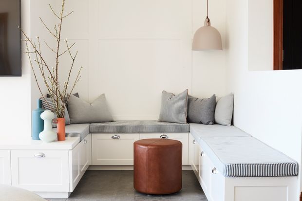 Bright built-in window-seat nook with white paneled cabinetry, striped upholstered bench cushions, neutral throw pillows, and a round brown leather ottoman. A soft pendant light, decorative branches, and a wall-mounted screen complete the clean, tailored interior.
