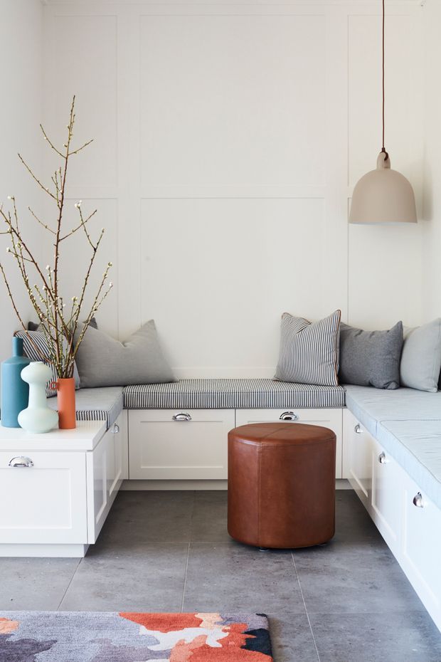 A bright, compact built-in window-seat or banquette area with white paneled walls and extensive white cabinetry beneath upholstered seating. The seating has striped cushions and soft grey pillows, with a large brown leather cylindrical ottoman in the centre. A simple pendant light hangs above, while colourful ceramic vases and bare branches provide decoration. Large grey floor tiles and a partially visible abstract rug complete the space.