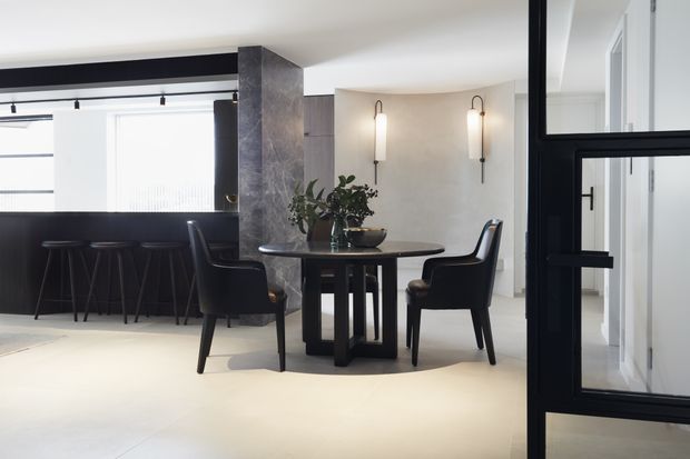 Refined contemporary dining area with a round dark dining table and upholstered chairs, positioned beside a black kitchen or bar counter with stools. The space features pale flooring and walls, dark framed glazing and doors, a textured dark stone-clad structural column, and slim wall sconces. A glazed black-framed doorway leads toward a bright adjoining area.