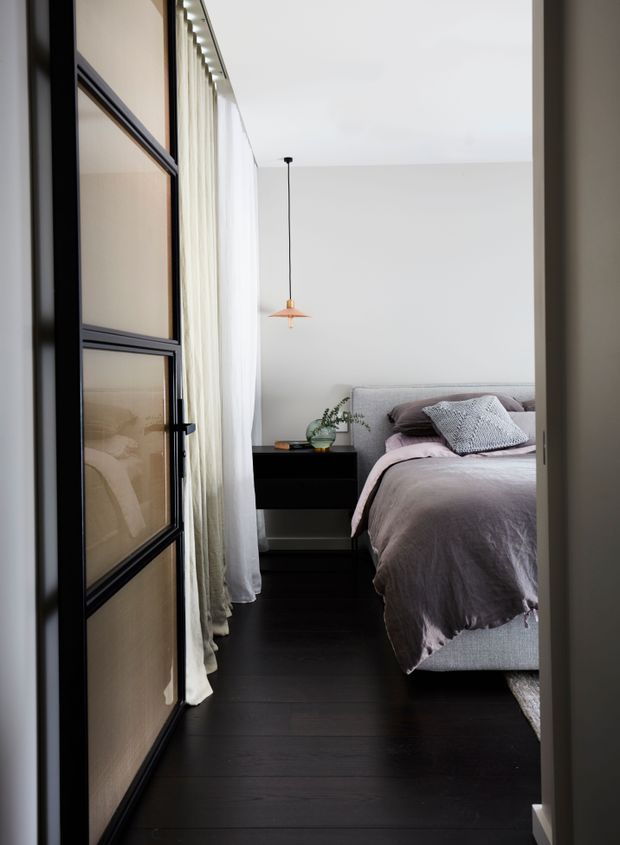 A compact, refined bedroom viewed through a partially open black-framed glazed door. The room features dark timber flooring, a low black bedside cabinet, a pale upholstered bed with soft grey-mauve bedding and textured cushions, sheer full-height curtains, and a simple copper pendant light. The palette is quiet and muted with strong contrast between the dark floor and pale walls and textiles.