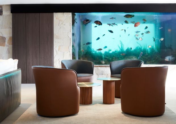 A sophisticated lounge area centered around a large built-in tropical aquarium. Four rounded brown leather lounge chairs and small wood-and-glass tables sit on a neutral rug, with dark vertical timber wall panelling and pale textured stone framing the aquarium.