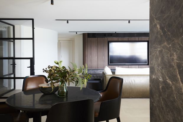 Refined open-plan dining and living area with a round dark stone dining table, upholstered dining chairs, sculptural greenery, and a cream sofa. A dark timber entertainment wall with a wall-mounted television sits beyond the dining space, while black-framed glazed partitions and a dramatic veined stone wall add architectural definition. Recessed and track lighting create a clean contemporary atmosphere.