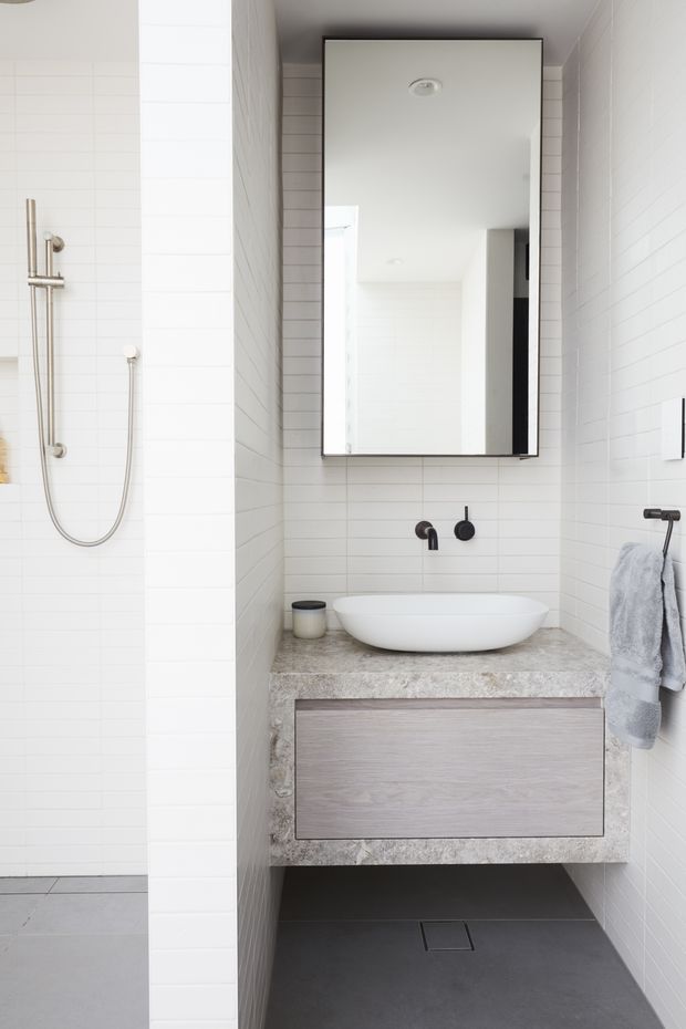 A bright, compact contemporary bathroom featuring white horizontal wall tiles, a dark grey tiled floor, a floating wood-grain vanity with a mottled stone benchtop, a white vessel basin, matte black tapware, and a large rectangular mirror. A tiled shower with exposed stainless-steel hardware is visible to the left.