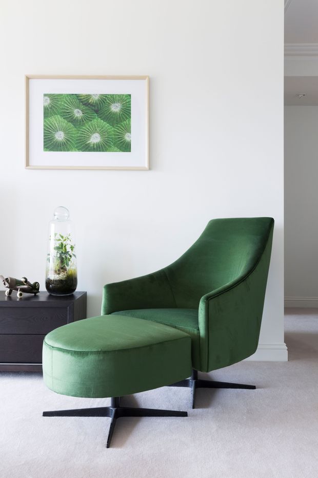 A refined interior vignette featuring a deep green upholstered lounge chair with a matching curved ottoman and black star-shaped base. The chair sits on a light neutral carpet beside a dark timber sideboard, with a glass terrarium and small sculpture on top. A framed botanical artwork hangs on the white wall, and a hallway or adjacent room is visible at right.