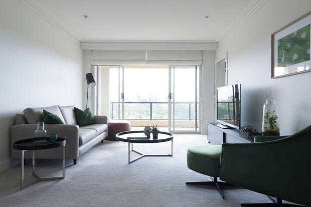 Bright contemporary living room with a neutral grey sofa, deep green accent armchair, round black coffee table, side table, television console, floor lamp and large glazed doors opening to a balcony. The room features white walls with decorative ceiling cornices, pale carpet and restrained modern furnishings.