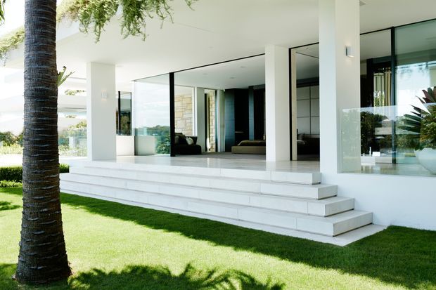 A bright contemporary home exterior with a deep covered terrace, broad white steps, slender white columns, and expansive floor-to-ceiling glazing. The open interior is visible through the glass, while a manicured lawn, palm trunk, and tropical planting frame the foreground. The design emphasizes clean geometric lines, strong indoor-outdoor connection, and a light neutral palette.
