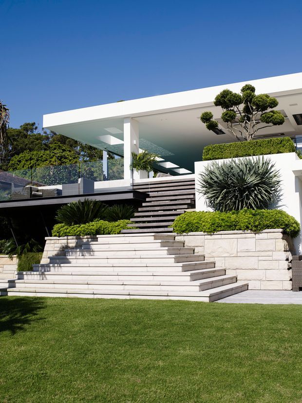 A bright exterior view of a modern multi-level residence with a flat-roofed white pavilion, expansive open terraces, broad stone steps, and landscaped garden beds. The facade combines white rendered surfaces, pale stone retaining walls and steps, dark horizontal decking or cladding, and glass balustrades. Sculpted topiary and architectural planting frame the entry terrace, with a manicured lawn in the foreground under a clear blue sky.