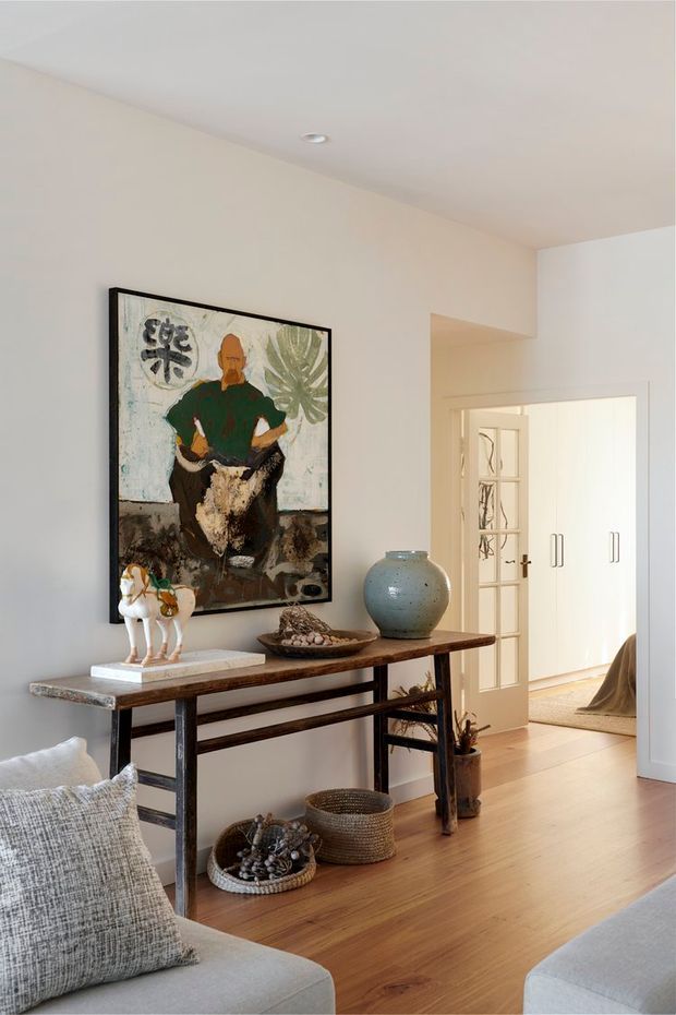 A warm, softly lit interior view of a living room or lounge area, featuring a slim rustic timber-and-metal console table against a pale wall, a large framed artwork, ceramic vessels, woven baskets, and a neutral upholstered sofa in the foreground. A doorway leads to a bright adjoining hallway or room with built-in white cabinetry. The room has light timber flooring, white walls and ceiling, and understated eclectic styling.