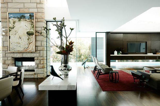 A spacious open-plan contemporary living and dining area with a dramatic stone-clad fireplace wall, dark timber flooring, a large white marble island or console, sculptural dining chairs, and a deep red rug. Full-height glazing opens the room to a leafy exterior, while a dark built-in media wall and linear fireplace define the far side of the space. The room is bright, refined, and strongly connected to the outdoors.
