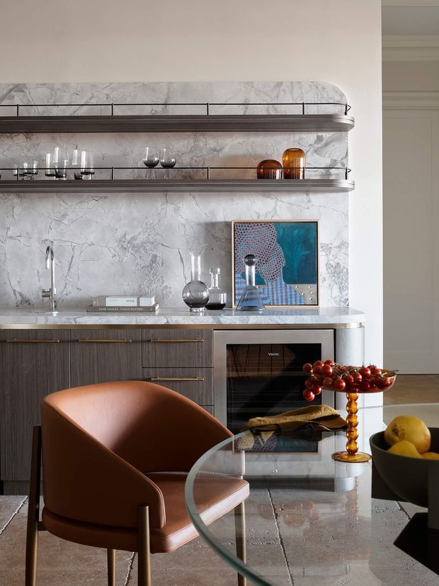 Refined contemporary kitchen and dining nook featuring a full-height veined marble backsplash, open display shelving, dark wood cabinetry, pale stone benchtop, integrated oven, and a sculptural brown leather dining chair around a glass table. Amber glassware, artwork, and decorative objects add warmth and colour.