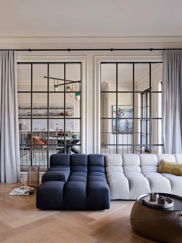 A refined living room with a large modular sectional sofa in dark navy and light grey upholstery, positioned on a pale herringbone timber floor. Full-height curtains frame black metal-framed glazed room dividers, revealing a sophisticated dining and bar area beyond. The room has high ceilings, ornate ceiling cornices, contemporary furniture, and a sculptural dark coffee table.