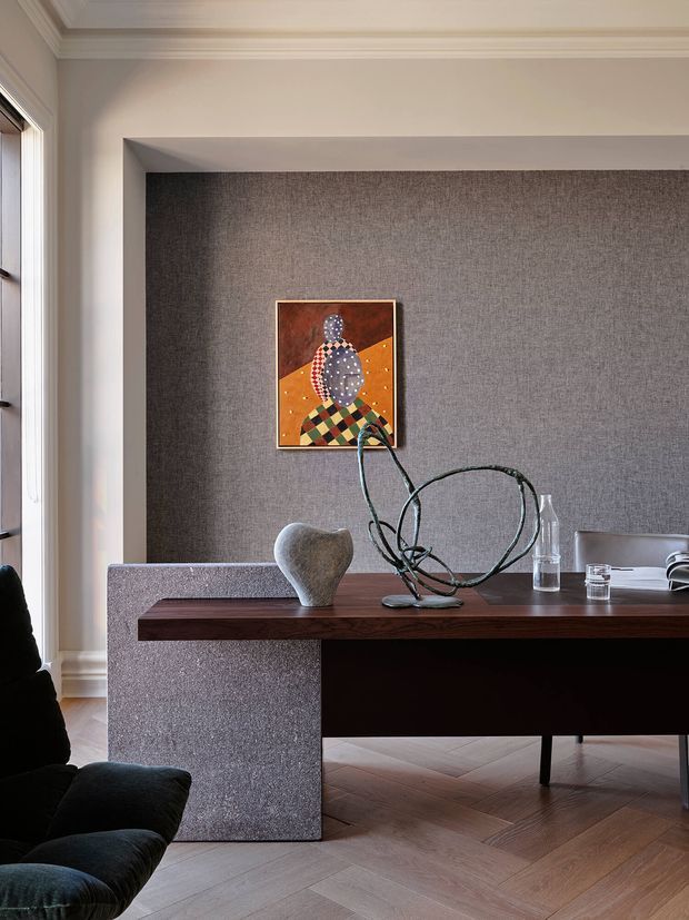 A refined interior vignette featuring a dark wood console or desk in front of a textured grey fabric-lined recessed wall. A black sculptural lounge chair, abstract artwork, ceramic vessel, metal sculpture, and glass objects add a curated, sophisticated feel. The room has warm herringbone timber flooring, pale walls, detailed traditional cornice moulding, and tall side glazing.