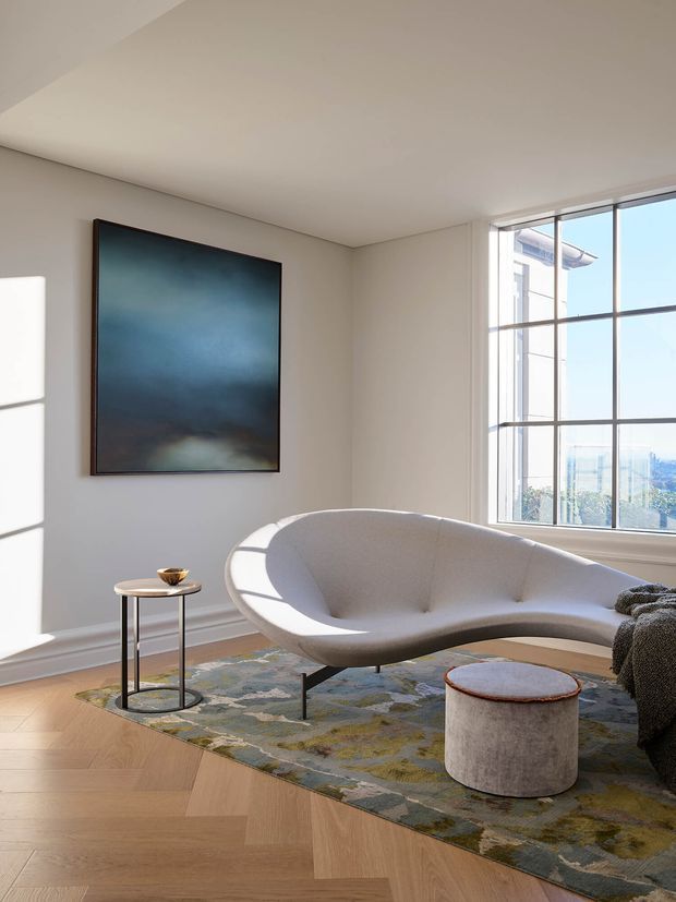A bright, refined living room with a sculptural cream curved sofa, small round side table, upholstered cylindrical ottoman, and abstract artwork. The room features pale walls with decorative lower moulding, a large black-grid window, warm herringbone timber flooring, and a muted patterned area rug. Strong natural light creates an airy contemporary atmosphere.