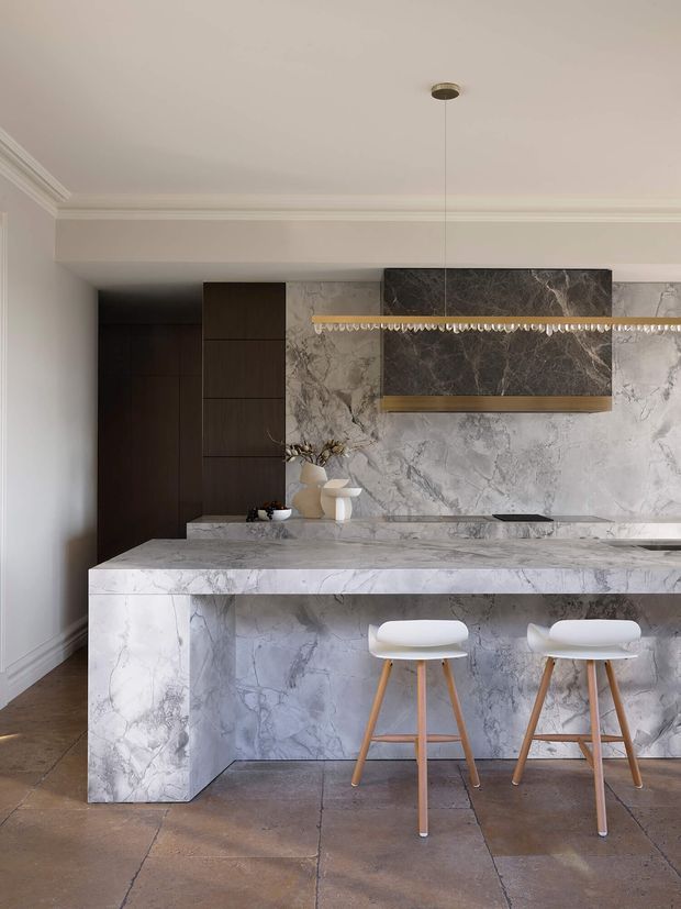 Refined contemporary kitchen featuring a large marble-clad island with waterfall ends and seating for two, dark timber cabinetry, a dramatic marble splashback, and a linear pendant light. Classic cornices and neutral tones contrast with the modern stone surfaces.