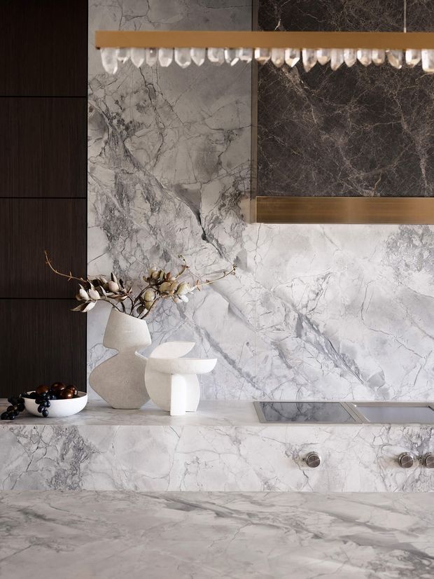 Close-up view of a luxurious stone-clad kitchen or wet bar, featuring dramatic white and grey veined marble across the backsplash, benchtop and lower cabinetry, dark timber cabinetry at left, and a bronze-framed mirrored or glass-fronted upper cabinet with decorative hanging glassware. Sculptural white ceramics and a fruit bowl sit on the counter.