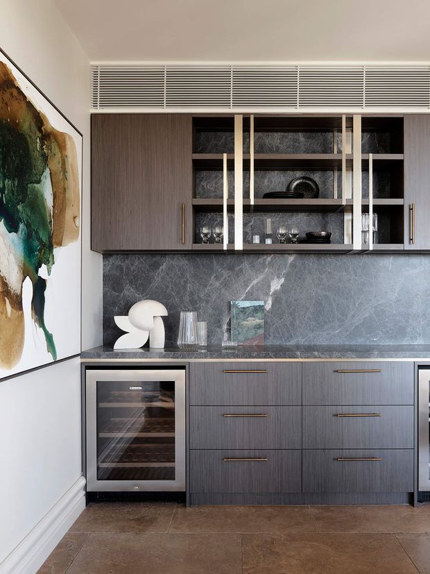 A refined contemporary kitchen or entertaining joinery wall featuring dark wood-grain cabinetry, a dramatic grey veined stone splashback and benchtop, integrated display shelving with glass-front doors, brass-toned handles, and a built-in wine or beverage cooler. A large abstract artwork and decorative objects add an artistic, luxurious character. The room has a tiled floor and a linear ventilation grille above the cabinetry.