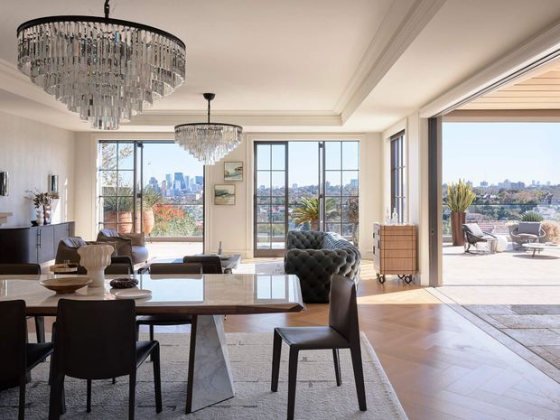 Elegant open-plan living and dining area with large black-framed windows and glazed doors opening to a sunlit terrace. The room features ornate crystal chandeliers, a marble-topped dining table, dark upholstered dining chairs, a tufted sofa, timber parquet flooring, decorative wall mouldings, and an expansive city view beyond.