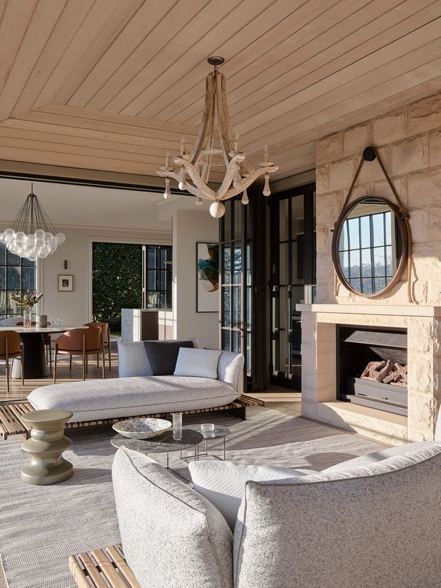 A refined living room with a large pale stone fireplace, black-framed glazed doors, and a light timber-lined coffered ceiling. Two curved light-grey sofas surround a round glass coffee table, while a sculptural antler-style chandelier and a spherical dining pendant add distinctive lighting. The room opens to a dining area with large windows and abundant natural light, creating a warm, sophisticated atmosphere.