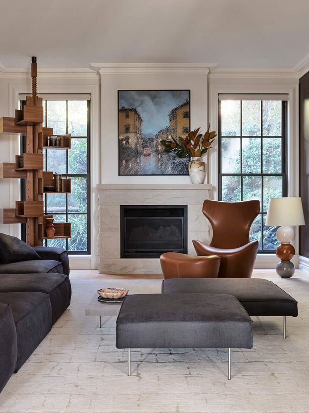 Refined living room with a large dark sectional sofa, matching ottoman, sculptural cognac leather lounge chair, pale stone fireplace, tall black-framed windows, traditional wall mouldings, and a timber open shelving unit. A muted architectural artwork and layered neutral finishes create a sophisticated, warm interior.