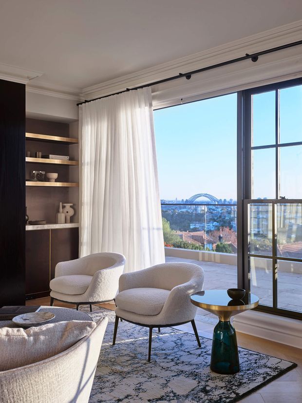 A refined living room with expansive black-framed windows and balcony access overlooking a city skyline and arched bridge. The room features sculptural cream boucle lounge chairs, a matching sofa, a patterned area rug, a small sculptural side table, sheer white curtains, dark built-in shelving with warm integrated lighting, and ornate ceiling cornices.