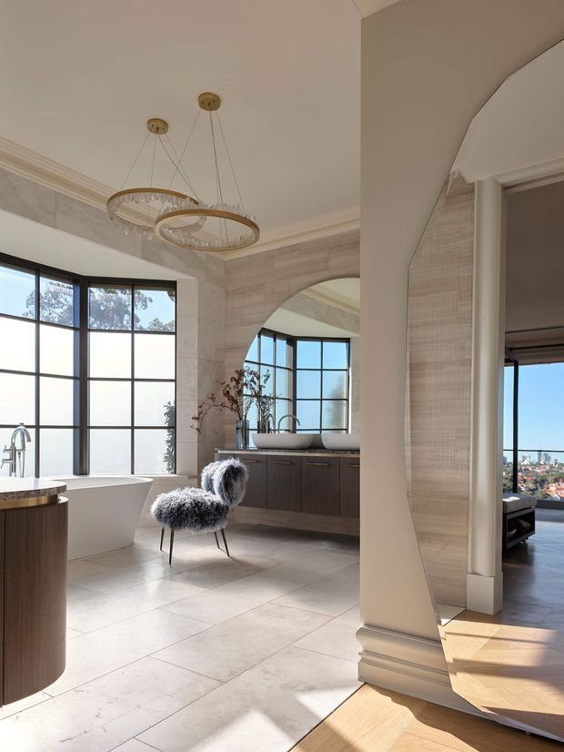 A bright, luxurious bathroom with a freestanding bathtub beside large black-framed windows, a dark timber vanity with twin vessel basins, pale stone-look tiled walls and floor, and an arched opening. Decorative pendant lighting, a textured accent wall, and a sculptural furry chair add refined character. A living area and city view are visible through the doorway.
