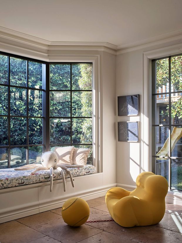 Sunlit, high-ceilinged interior with ornate layered cornice mouldings, tall black-framed multi-pane windows, and a deep built-in window seat. The room features pale walls, a tiled floor, a patterned upholstered cushion, framed artwork, and sculptural mustard-yellow seating and ottoman, creating a playful yet refined setting with strong garden views.