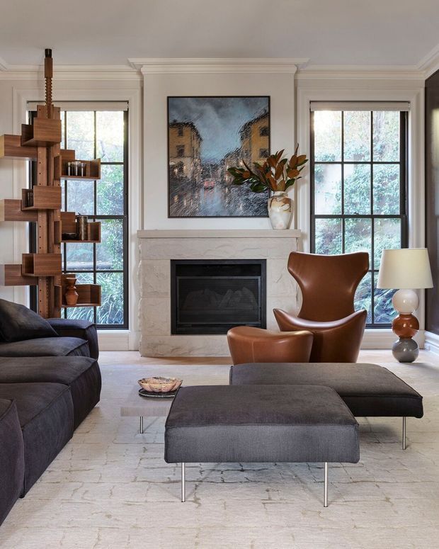 Refined living room with a central white stone fireplace, tall black-framed windows, a dark sectional sofa, brown leather lounge chair, matching ottoman, and a sculptural timber open shelving unit. Neutral finishes, classic wall mouldings, and contemporary furnishings create a warm, sophisticated interior.